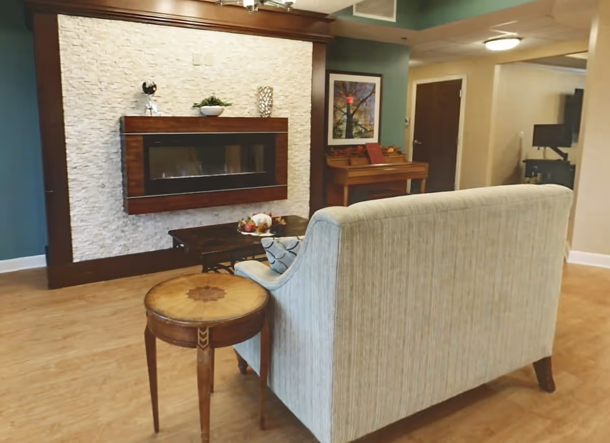 A cozy living room area in an assisted living facility featuring a light-colored upholstered sofa, a round wooden side table, a dark wooden coffee table with decorative items, and a modern wall-mounted fireplace set against a textured white stone wall. There is a framed picture on the wall and a wooden piano in the background near a closed door. The room has wooden flooring and soft lighting.