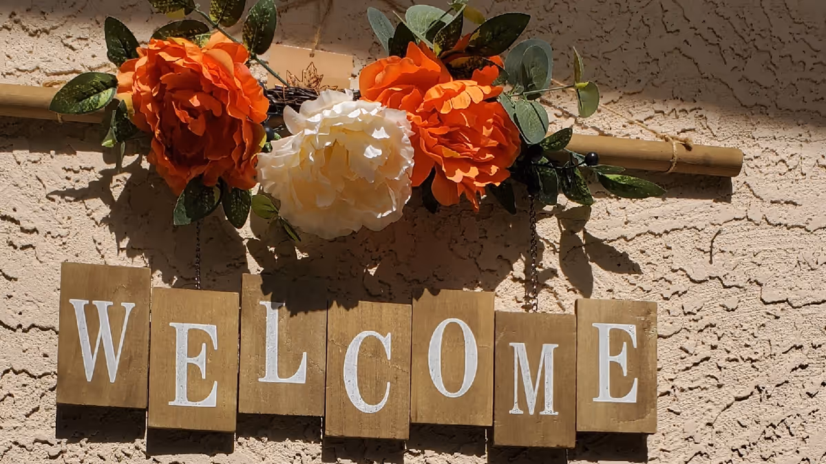 Wooden 'WELCOME' sign with orange and white artificial flowers hanging on a textured exterior wall.