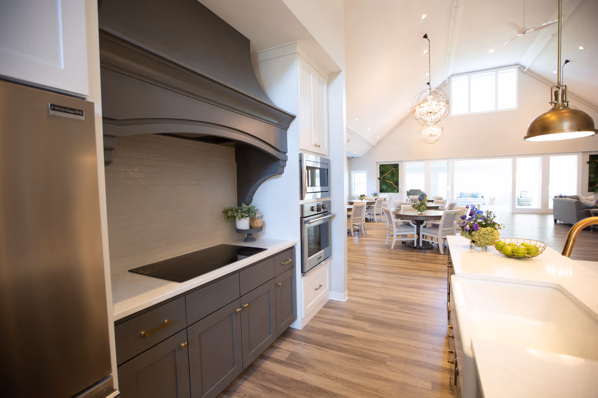 Bright and spacious kitchen area with modern appliances including a stainless steel refrigerator, built-in oven, and stovetop under a large dark range hood. The kitchen features white countertops, gray lower cabinets, and a white sink with a brass faucet. Beyond the kitchen, there is a dining area with round tables and chairs, decorated with floral centerpieces. The space has high vaulted ceilings with large windows allowing natural light to fill the room.
