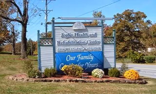 Outdoor view of a sign for Joplin Health and Rehabilitation Center located at 221 S.W. 32nd St. The sign includes the center's phone number and a blue banner that reads 'Our Family Caring for Yours...' surrounded by landscaping with flowers and trees in the background.
