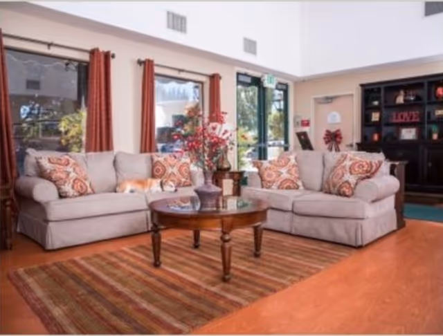 Communal lounge with two beige sofas facing a round wooden coffee table on a striped rug, decorative pillows and shelving.
