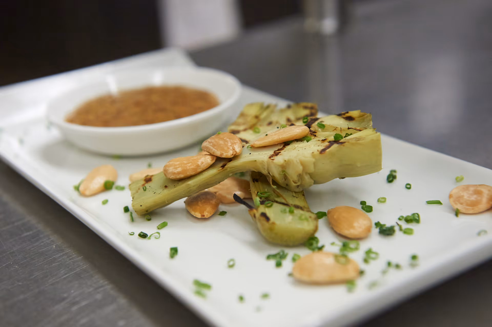 A close-up of a white rectangular plate with grilled artichoke halves garnished with chopped chives and several almonds, accompanied by a small white bowl of dipping sauce in the background.
