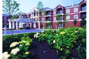 Exterior view of a multi-story senior living facility named AdamsPlace, featuring a red brick facade, white columns at the entrance, and landscaped greenery with bushes and flowers in the foreground.