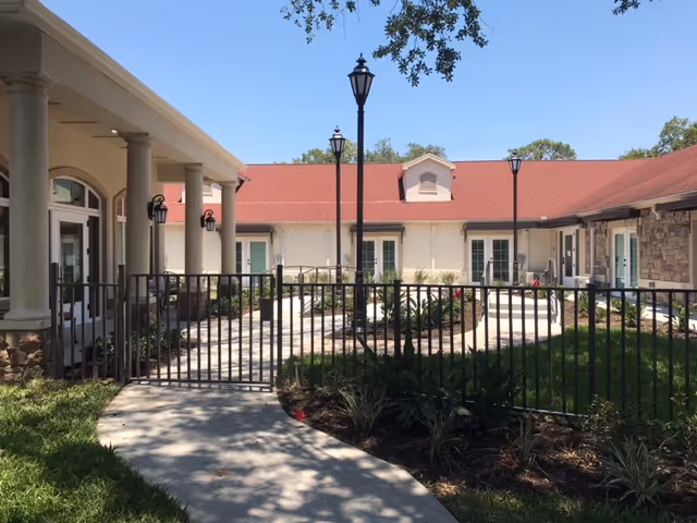 Outdoor courtyard area of a senior living facility with a paved walkway, black metal fence, green grass, plants, and a building with a red roof and white walls under a clear blue sky.