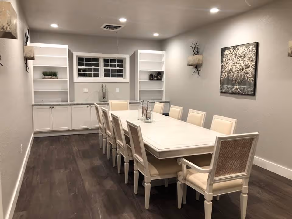 A modern dining room with a long white rectangular table surrounded by ten beige cushioned chairs. The room features dark wood flooring, white built-in shelves and cabinets along the back wall, a small window above the cabinets, and neutral-colored walls adorned with a decorative wall art and two wall sconces.