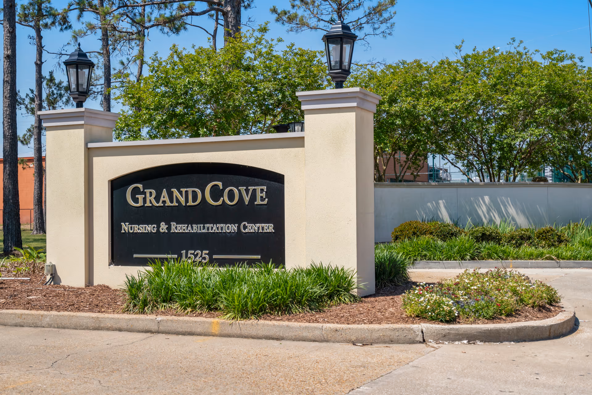 Entrance sign for Grand Cove Nursing & Rehabilitation Center surrounded by landscaping with green bushes and trees in the background under a clear blue sky.