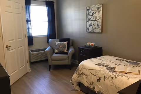 A small bedroom with a bed covered in floral bedding, an open book on the bed, a cushioned armchair with a decorative pillow, a window with dark curtains, a nightstand with a box of tissues, and a wall painting featuring birds and branches.