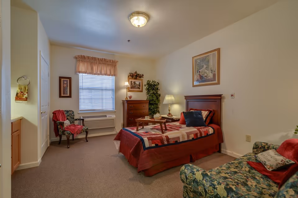 Well-lit furnished bedroom with a single bed, dresser, armchairs, and a window with blinds.