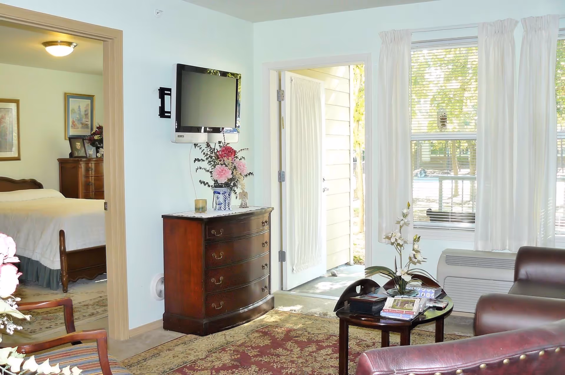 A bright living room with a leather armchair and a small round table holding books and a flower vase. A flat-screen TV is mounted on the wall above a wooden chest of drawers with a floral arrangement. The room has a large window with white curtains and a door leading outside. A bedroom with a bed and dresser is visible through an open doorway.