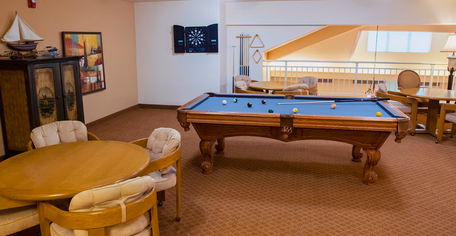 Recreation room with a pool table, round tables and cushioned chairs, a dartboard on the wall, and warm carpeting.