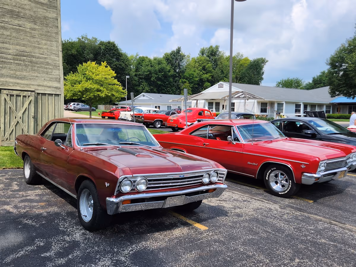 Two vintage red cars parked side by side in a parking lot with a building and trees in the background under a partly cloudy sky.