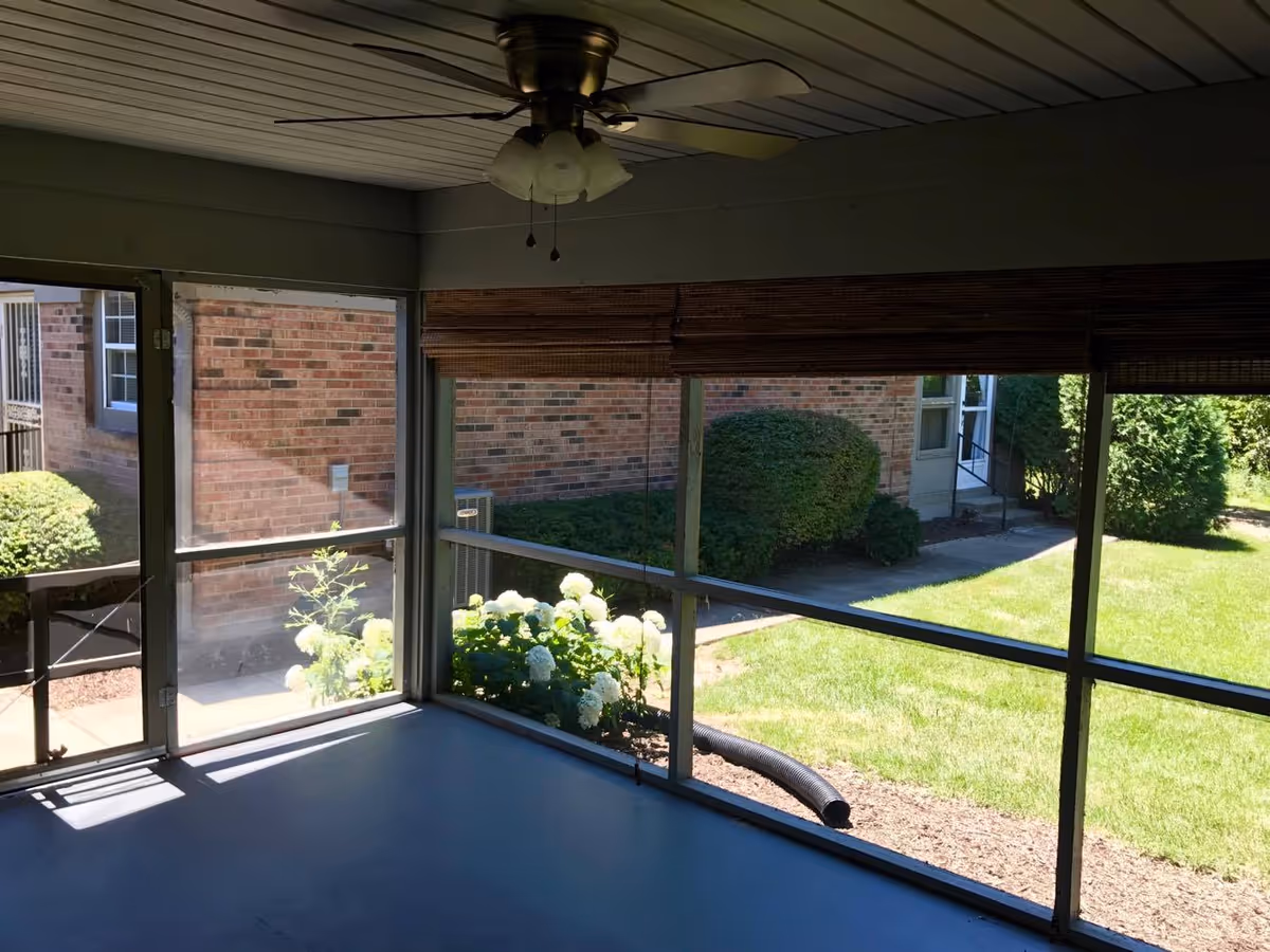 View from a screened-in porch showing a ceiling fan with lights, a door leading outside, and a garden area with white flowers and green bushes. Beyond the porch, there is a grassy lawn and brick building walls.