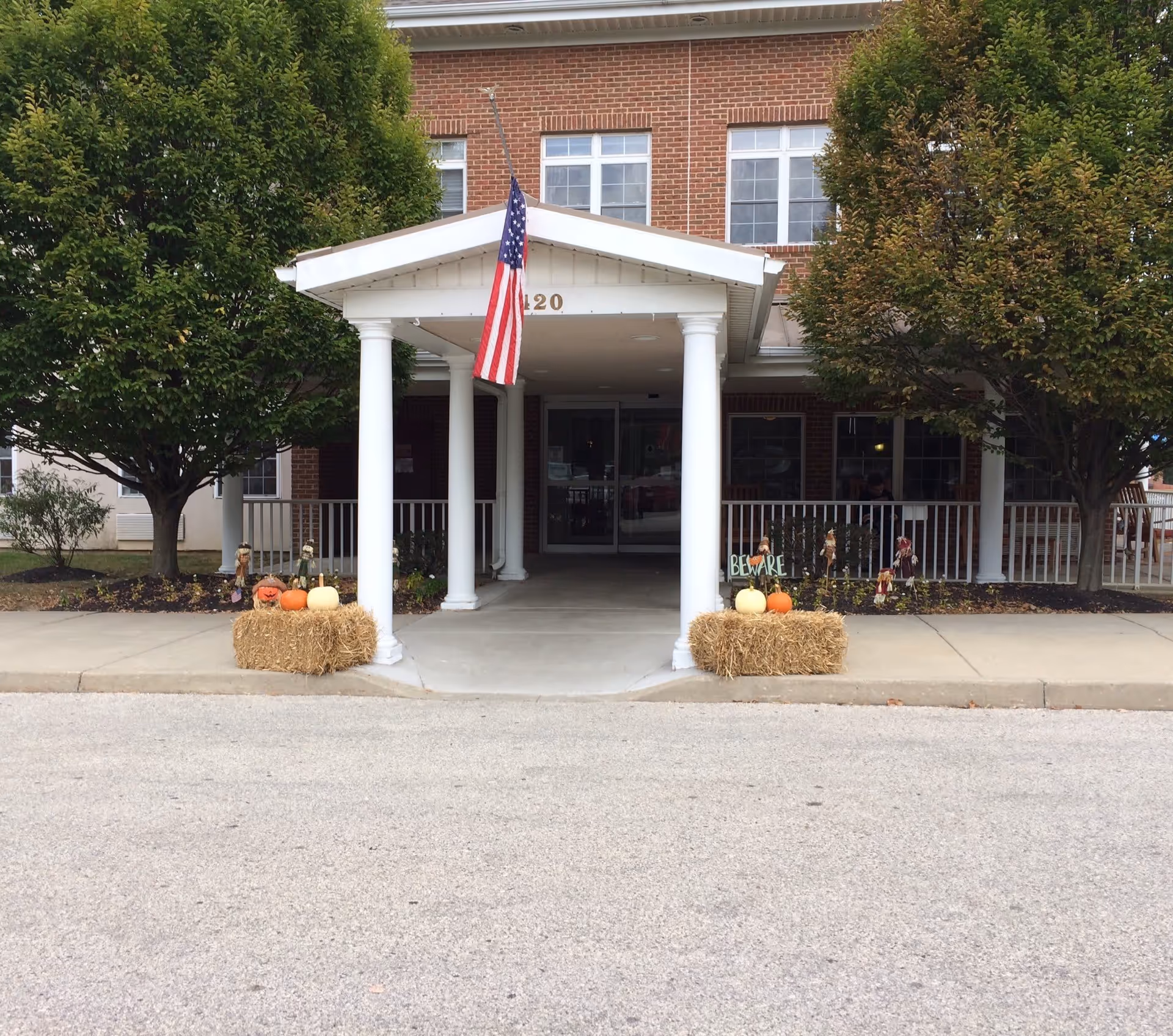 Front entrance of a brick building with white columns supporting a small roof over the walkway. An American flag hangs from the roof. Two trees flank the entrance, and hay bales decorated with pumpkins and scarecrows are placed on either side of the walkway. The building has multiple windows and a glass door entrance.