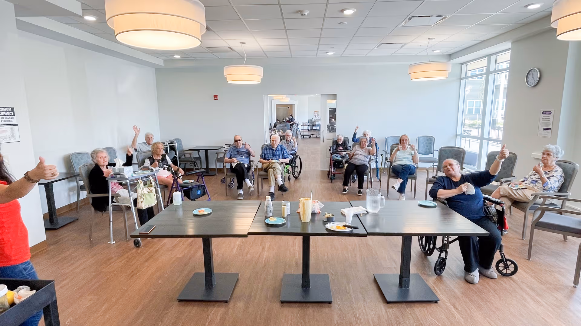 A group of elderly residents seated in a bright communal dining/activity room, many giving thumbs up.