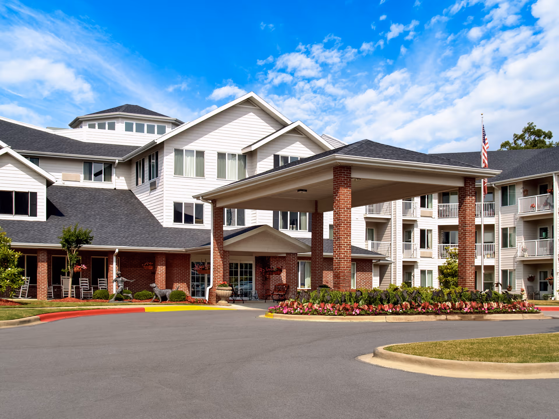 Exterior view of Asher Point Independent Living of Hoover showing a large building with white siding and brick accents, a covered entrance supported by brick columns, landscaped flower beds, and a clear blue sky with some clouds.