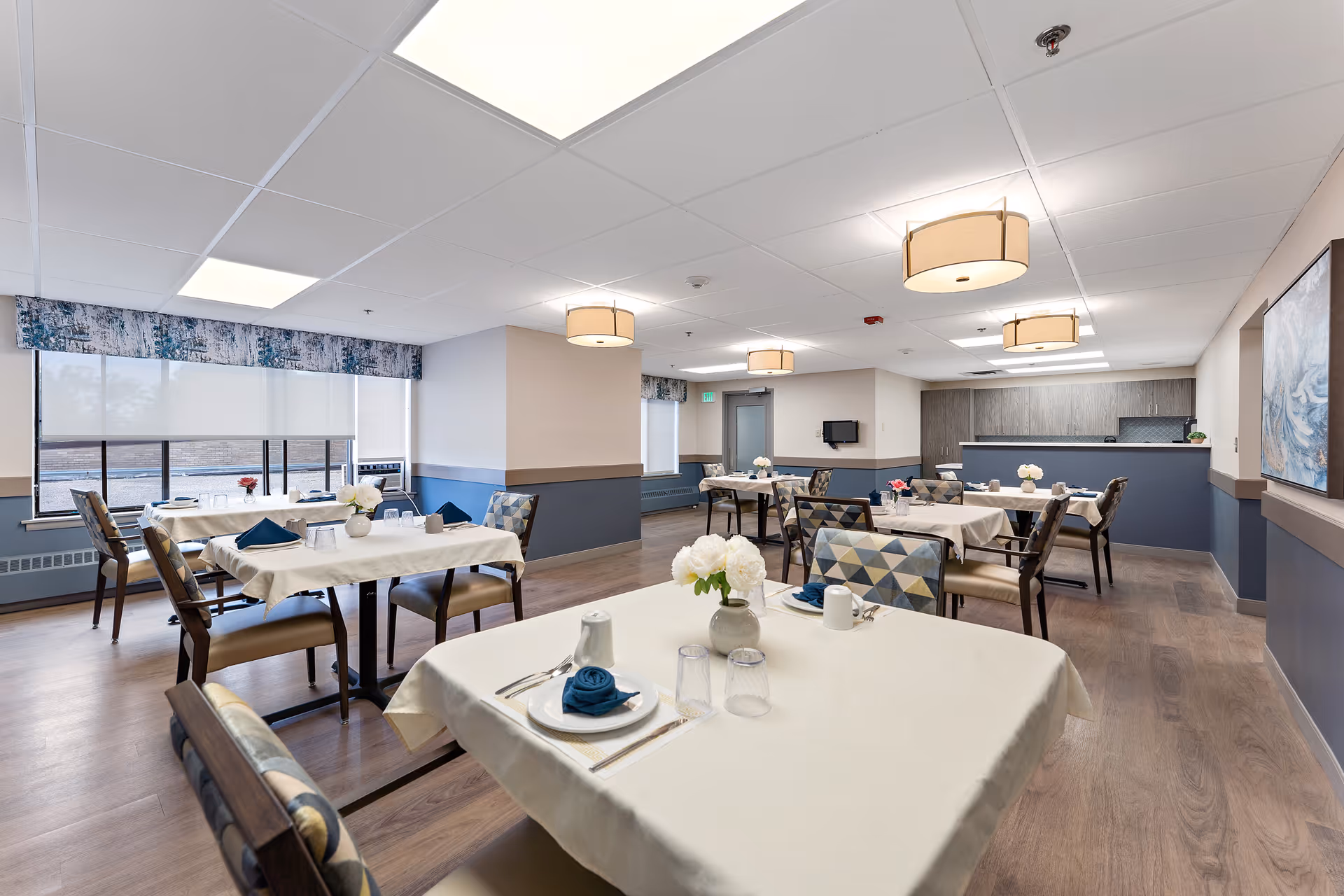 Spacious, well-lit dining room with multiple tables set for meals, upholstered chairs, and overhead pendant lights.