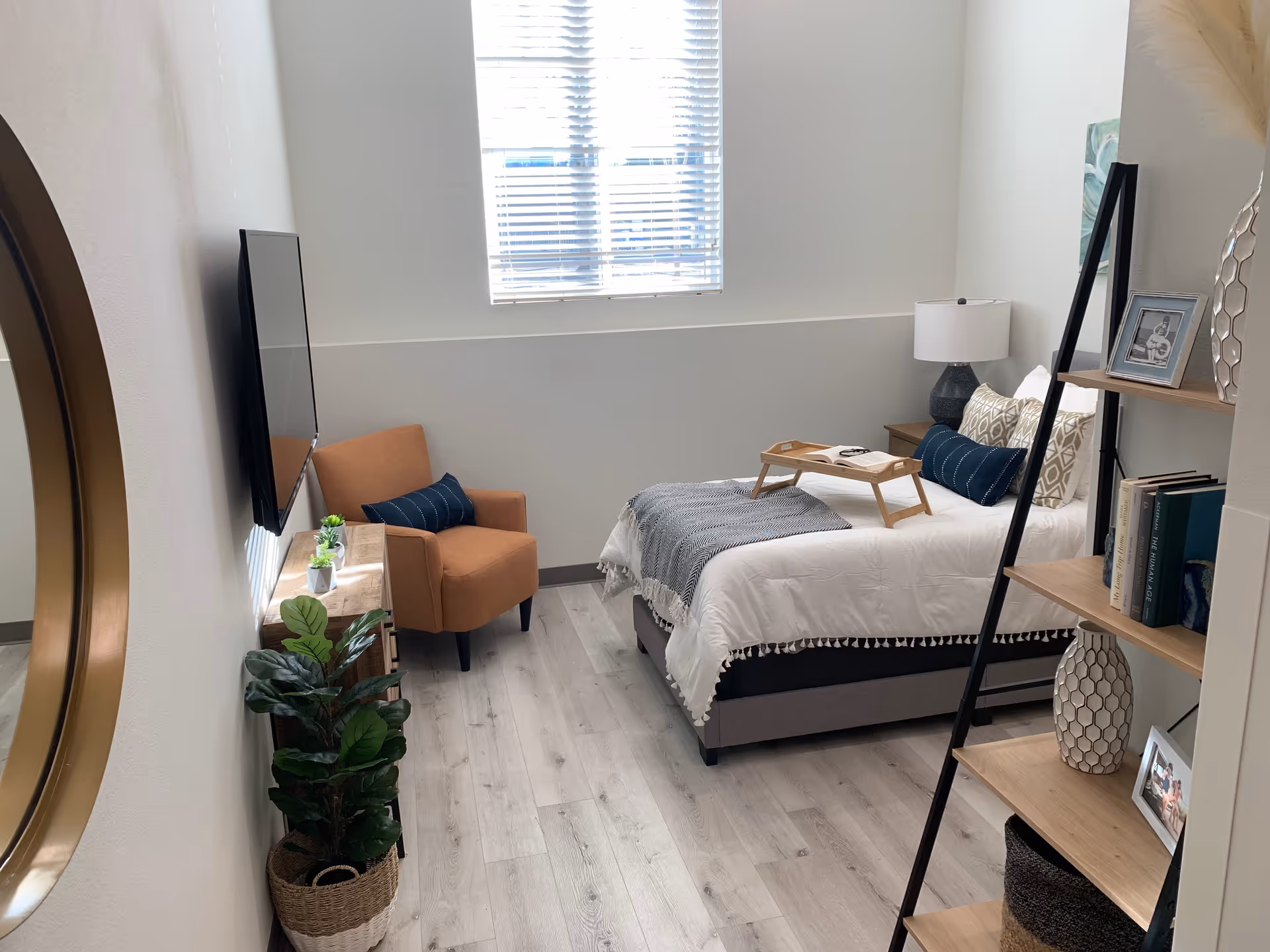 A bright and cozy bedroom with a bed dressed in white and gray linens, a wooden tray with a book on the bed, an orange armchair with a dark blue pillow, a wall-mounted flat-screen TV, a tall black and wood bookshelf with decorative items and books, a potted plant in a woven basket, and a window with blinds letting in natural light.