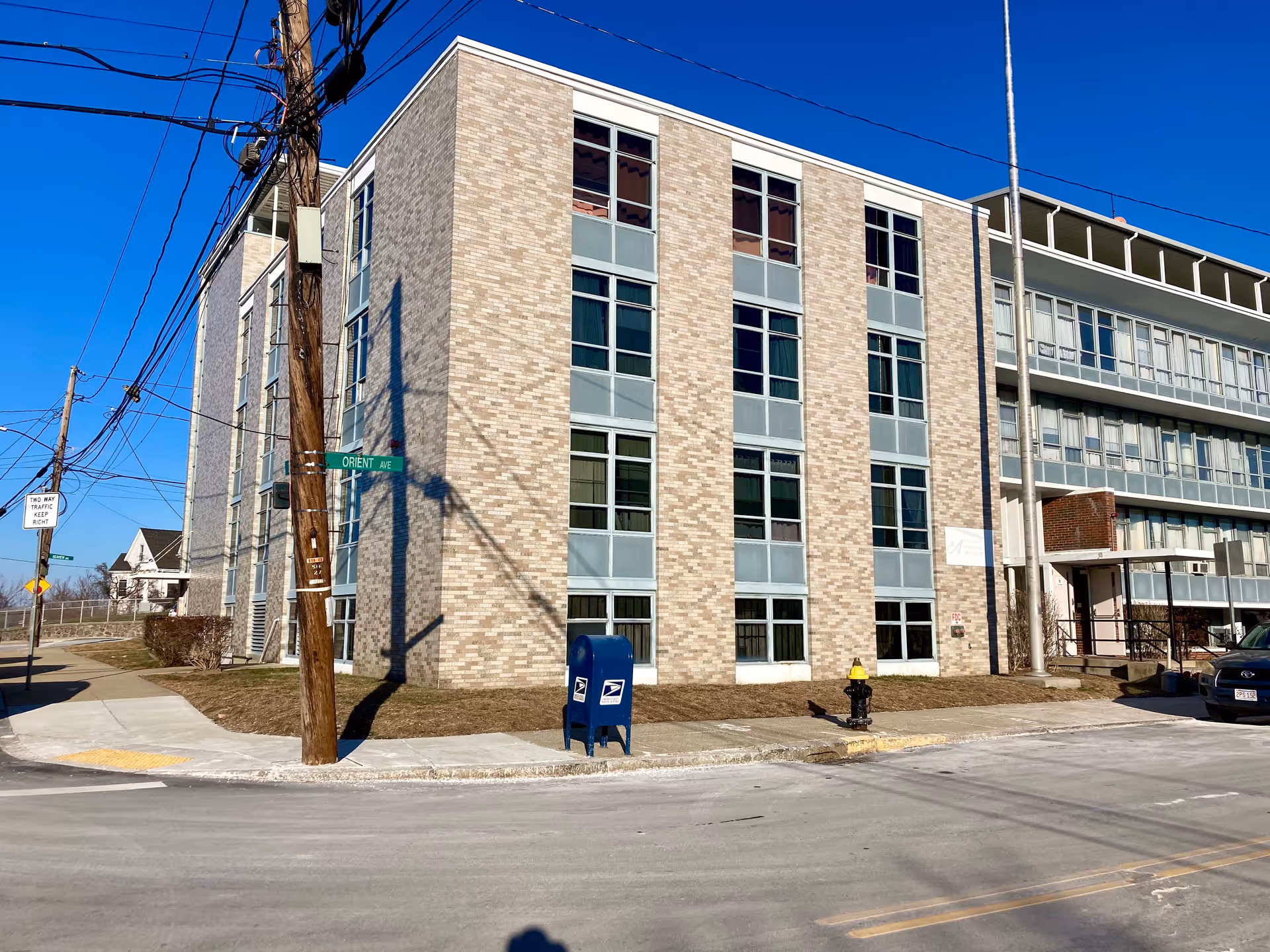 Exterior view of a multi-story brick building on a street corner with a blue mailbox and a fire hydrant on the sidewalk. There is a street sign labeled Orient Ave and a utility pole with wires. The sky is clear and blue.