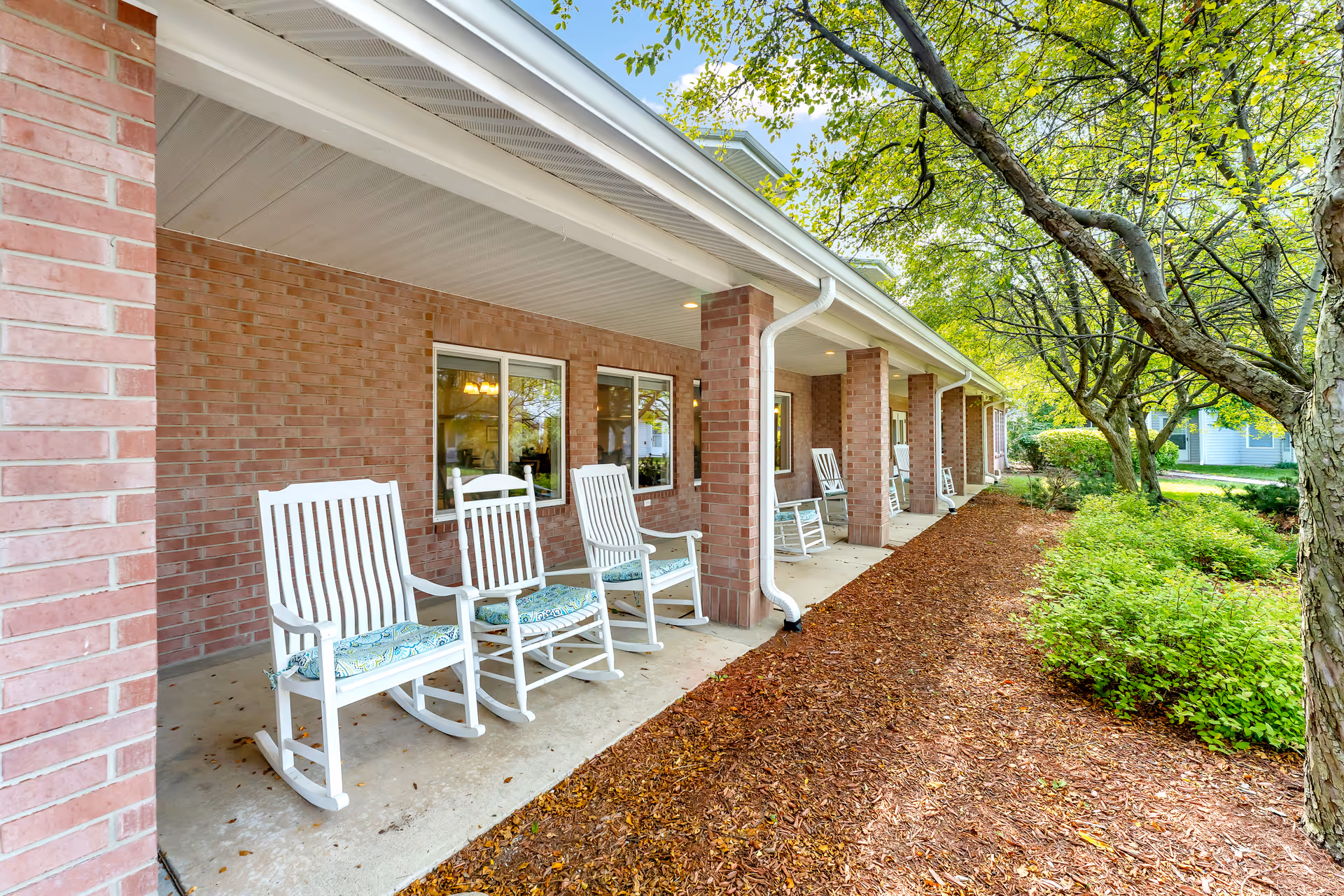Covered brick porch with white rocking chairs along a landscaped walkway under trees.
