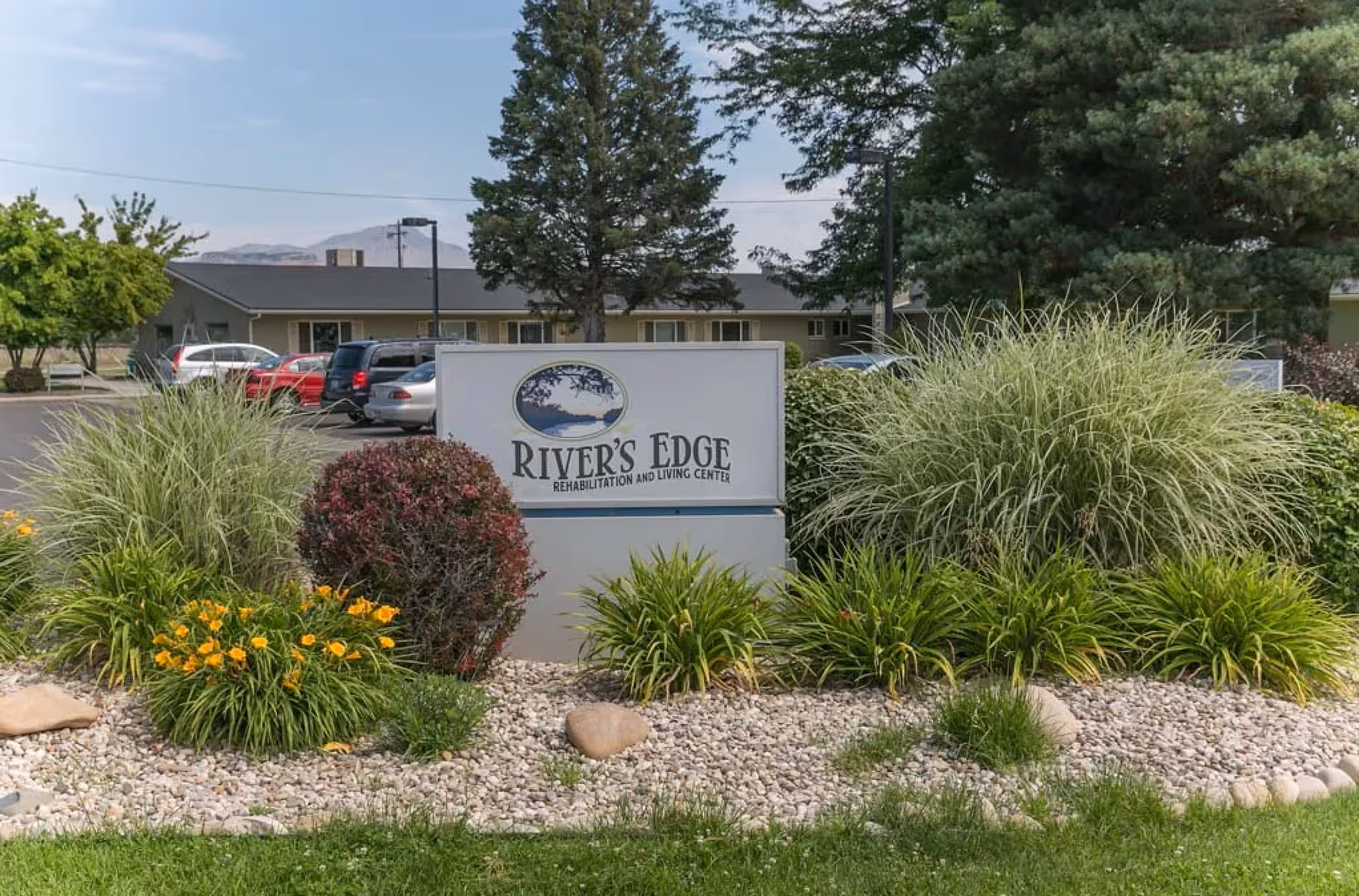 Outdoor view of River's Edge Rehabilitation and Living Center sign surrounded by landscaped bushes, flowers, and rocks with a building and parked cars in the background under a clear sky.