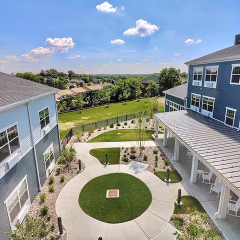 View of an outdoor courtyard area between two blue multi-story buildings with white trim, featuring a circular concrete walkway with a grass center, landscaped plants, and a covered patio with white chairs under a clear blue sky with scattered clouds.