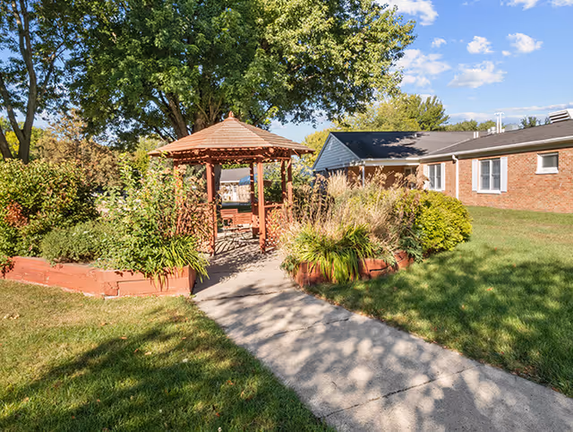 A sunny outdoor garden area with a wooden gazebo surrounded by lush green plants and trees. A concrete pathway leads to the gazebo, and a brick building is visible in the background under a blue sky with scattered clouds.