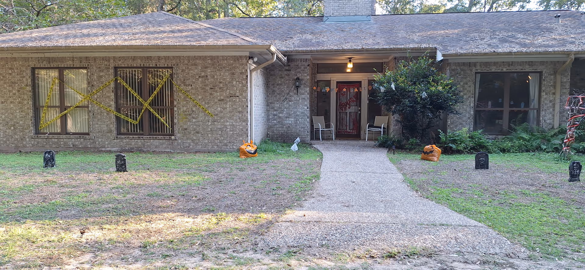 Front exterior view of a single-story brick house with a shingled roof. The house has two large windows on either side of a central entrance with a covered porch. The windows on the left are decorated with yellow tape in a crisscross pattern. The porch has two chairs and a door decorated with Halloween-themed items. The front yard features a gravel walkway leading to the porch, with Halloween decorations including small tombstones and pumpkin bags placed on the grass.