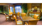 A cozy interior room with multiple wooden tables and cushioned chairs arranged for seating. The room features large windows with blinds, green plants, and a decorative cabinet with a vintage gramophone on top. The walls have framed artwork and the carpet is a muted red color.