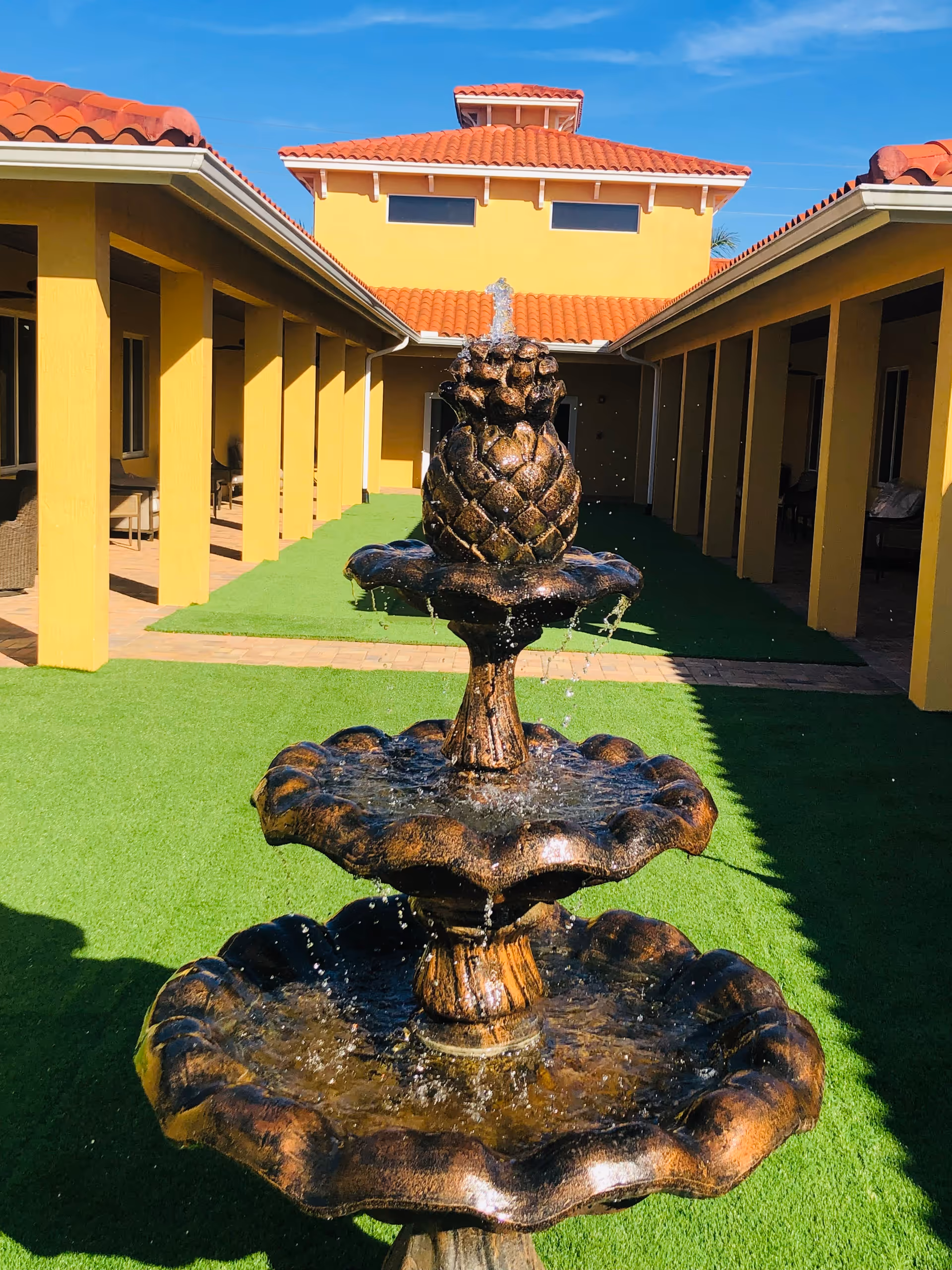 Three-tiered decorative fountain in a sunny courtyard flanked by yellow buildings with red tile roofs and artificial grass.