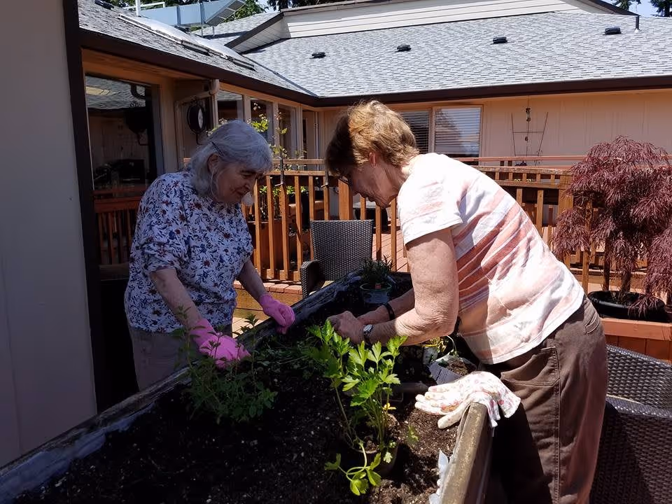 Two elderly women gardening together on a raised garden bed on a sunny outdoor patio with wooden railings and chairs nearby.
