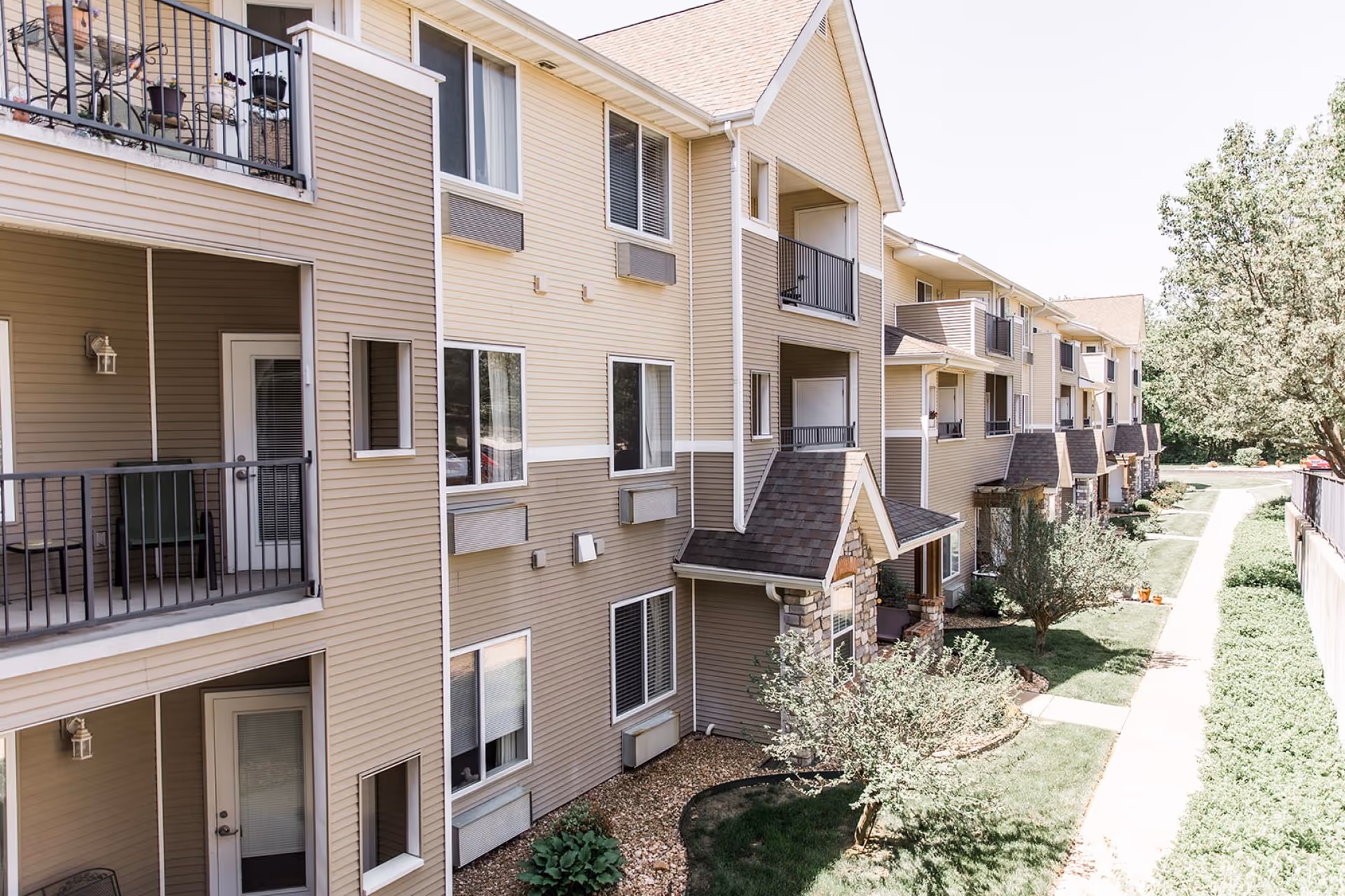 Exterior view of a multi-story residential building with beige siding and balconies. There is a walkway alongside the building with grass, small trees, and shrubs. The sky is clear and the area appears well-maintained.