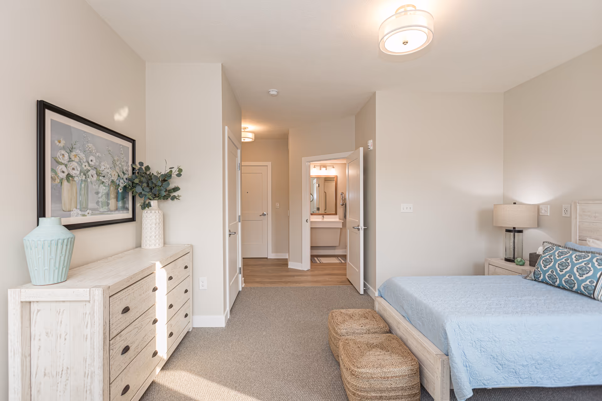 A bright and clean bedroom featuring a light wood dresser with decorative vases and a framed floral painting above it. The room has a bed with a light blue quilt and patterned pillows, a nightstand with a lamp, and two woven ottomans at the foot of the bed. An open door reveals a bathroom with a sink and mirror. The walls are painted light beige and the floor is carpeted with a wood floor visible near the bathroom and hallway.
