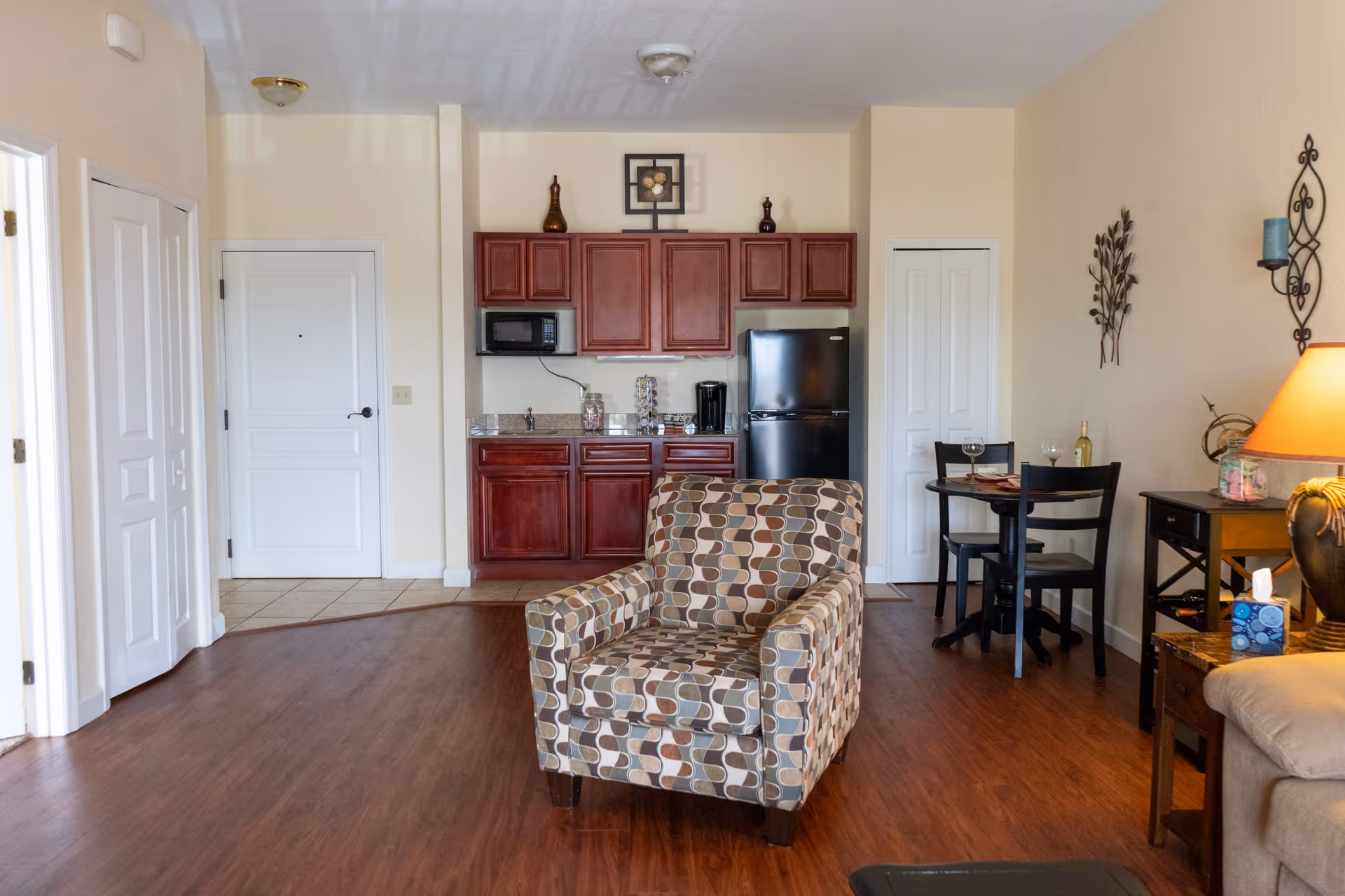 Interior view of a senior living facility room at Sunflower Springs featuring a patterned armchair in the foreground, a small kitchen area with wooden cabinets, a black refrigerator, microwave, and coffee maker in the background, and a small dining table with two chairs to the right. The room has wooden flooring, beige walls, and decorative wall art and a lamp on a side table.