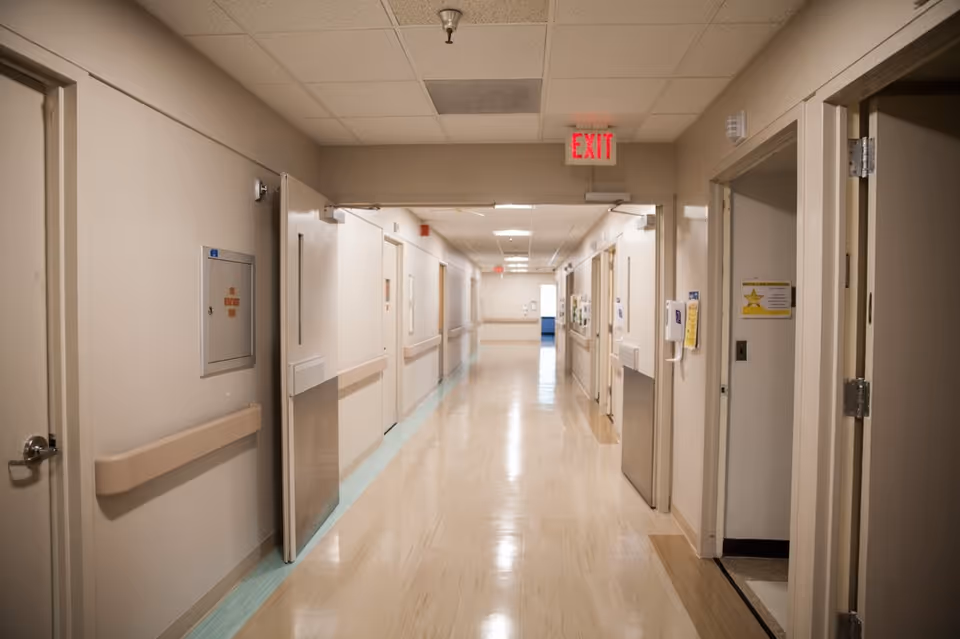 A clean, well-lit hospital hallway with beige walls and light-colored flooring. Doors line both sides of the corridor, some slightly open, and handrails run along the walls. An illuminated red EXIT sign is visible on the ceiling near the center of the image.