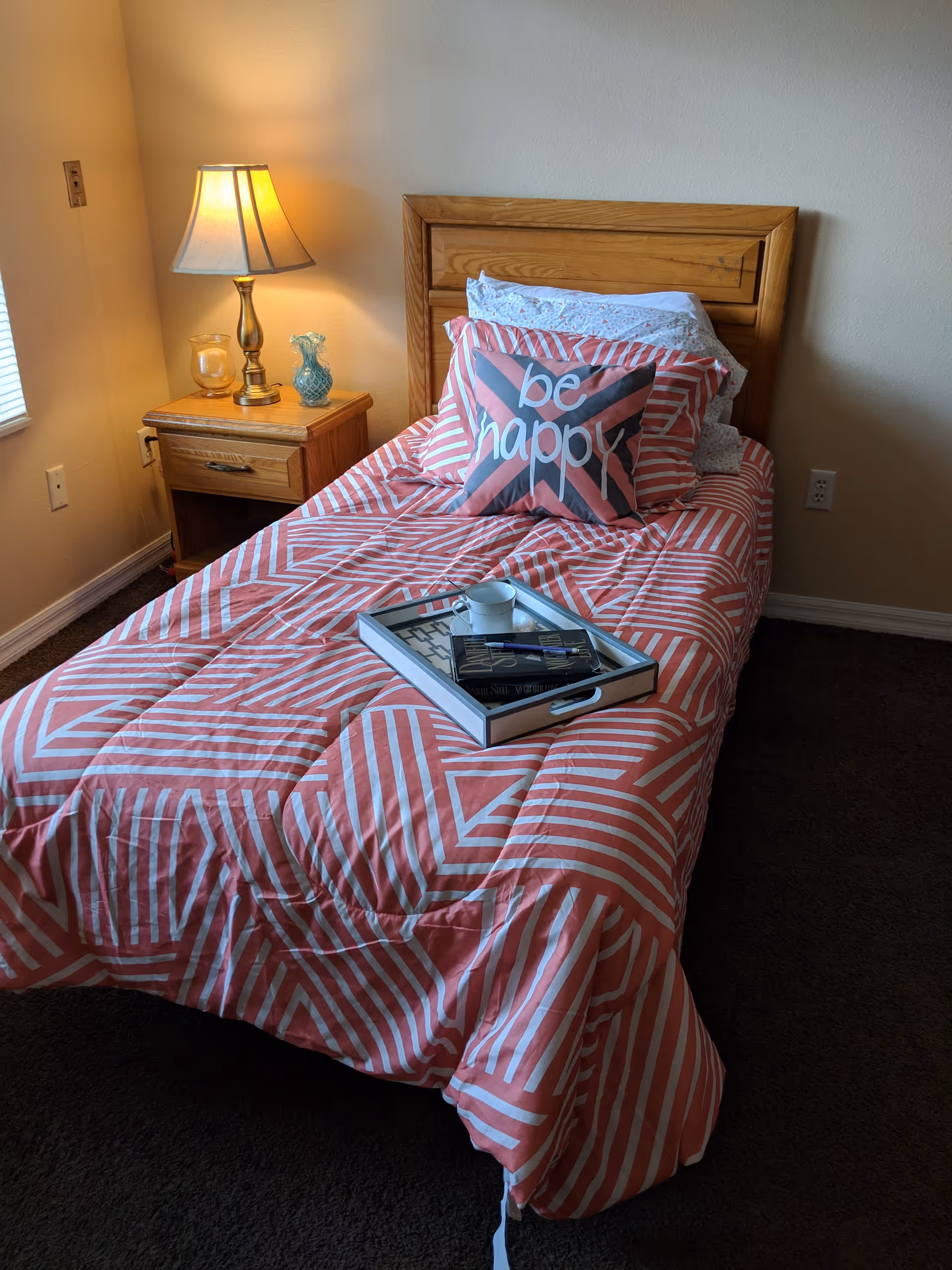A single bed with a wooden headboard is made up with a red and white geometric patterned comforter and pillows, including one with the words 'be happy'. A tray with a cup, book, and pen rests on the bed. Next to the bed is a wooden nightstand with a lit table lamp, a candle, and a decorative glass item. The room has beige walls and a window with blinds partially visible on the left.
