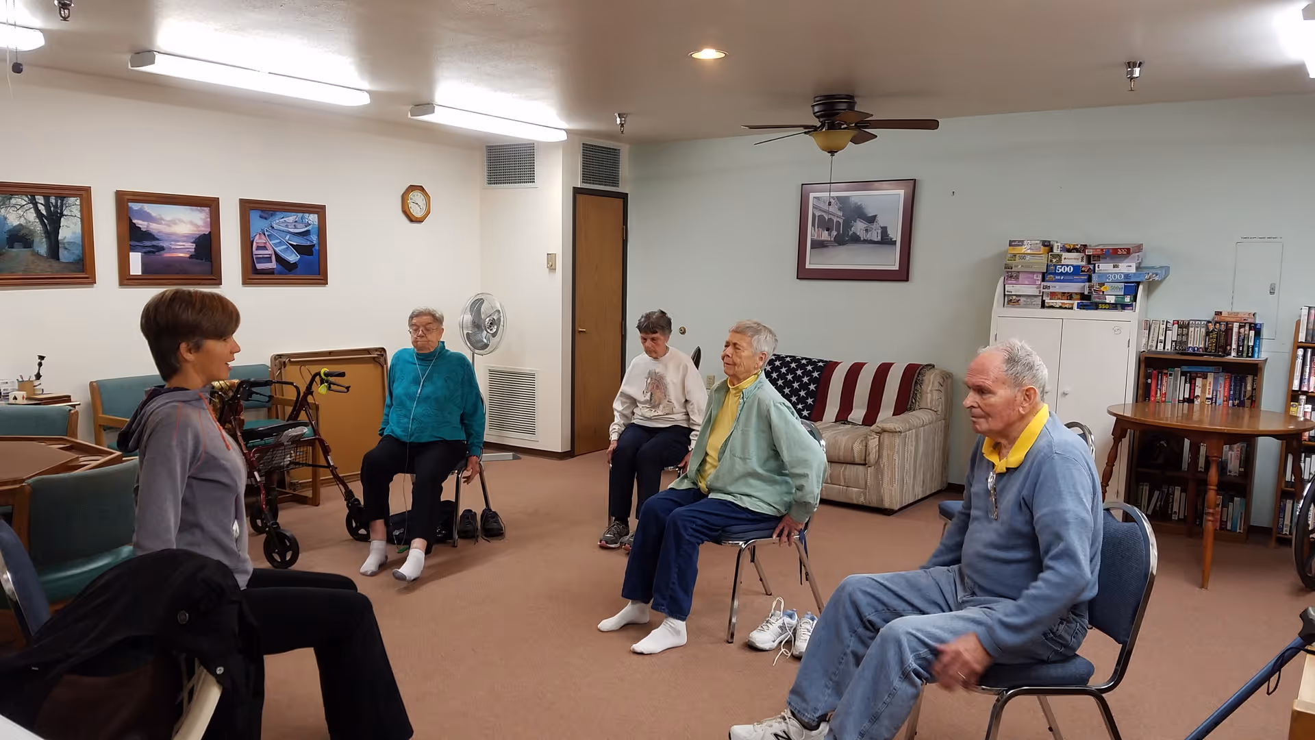 A group of elderly individuals seated on chairs in a common room participating in a seated exercise session led by an instructor. The room has framed pictures on the walls, a ceiling fan, a couch with an American flag blanket, bookshelves, and board games stacked on a cabinet.