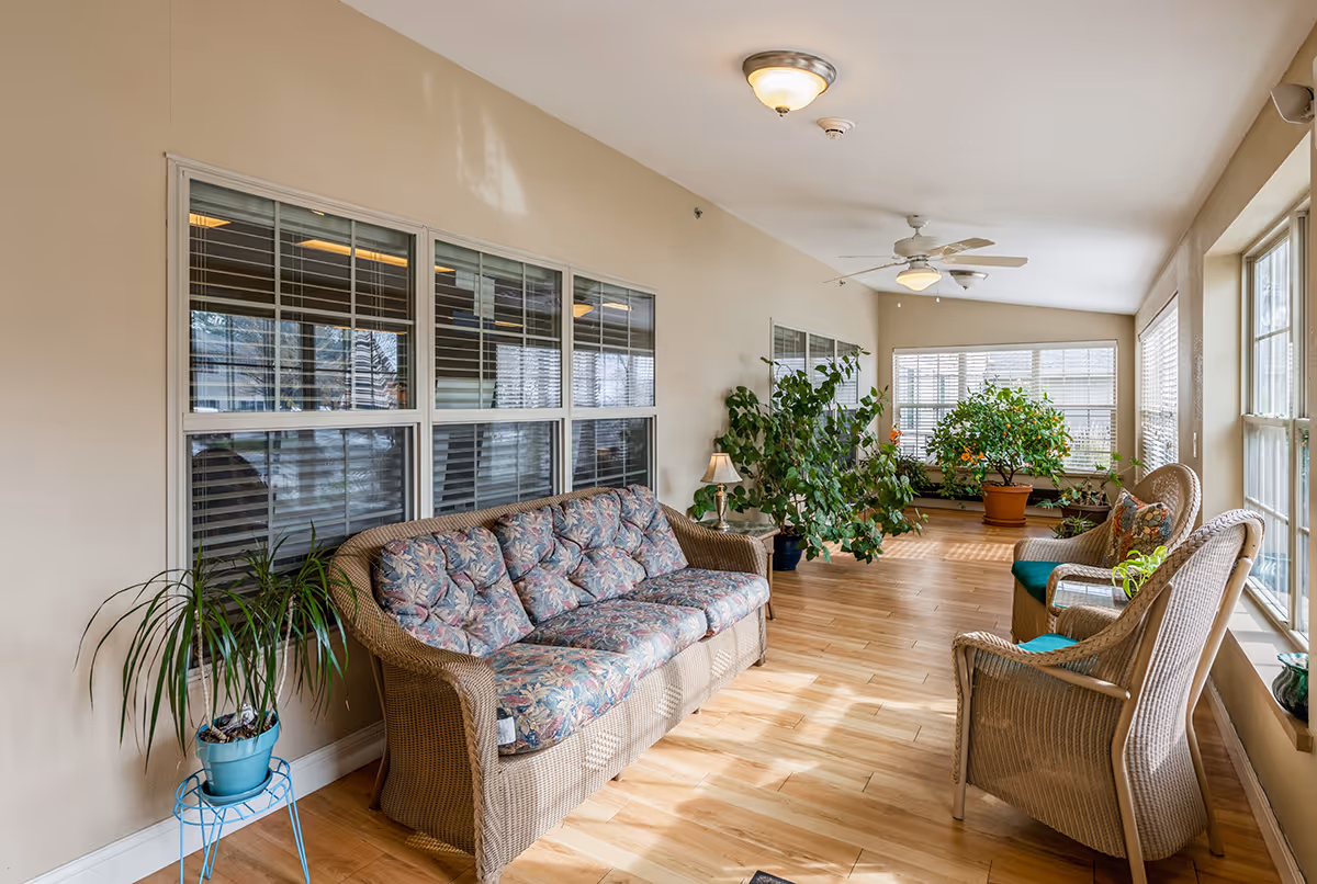 Sunlit indoor sitting area with wicker sofa and chairs, potted plants, and large windows.