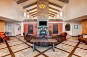 Spacious assisted living facility common area with high vaulted ceiling featuring exposed wooden beams, a large stone fireplace with a mounted flat-screen TV, and comfortable seating arranged around the fireplace. The floor has a tiled pattern with dark borders, and there are decorative plants and framed artwork on the walls.
