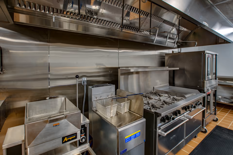A commercial kitchen with stainless steel appliances including a deep fryer with baskets, a large stove with multiple burners, an oven, and a stainless steel backsplash and ventilation hood.