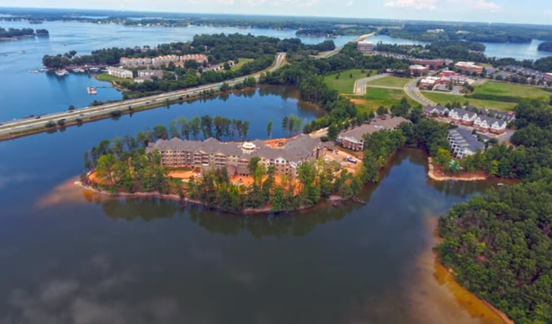 Aerial view of a retirement living facility named Williams Place Gracious Retirement Living situated on a peninsula surrounded by water, with trees and greenery around the buildings. Nearby roads, other residential buildings, and a large body of water are visible in the background under a partly cloudy sky.