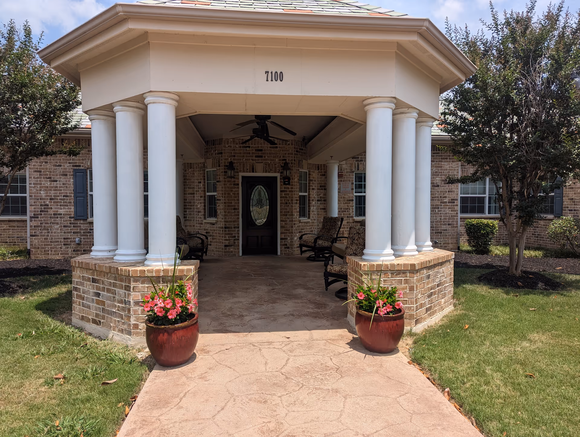 Entrance to a brick building with a covered porch supported by white columns. Two large flower pots with pink flowers flank the walkway leading to a door with decorative glass. The building number 7100 is displayed above the porch.