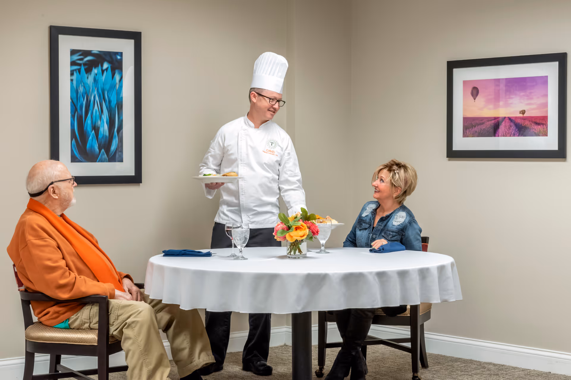 A chef wearing a white uniform and hat serves food to an elderly man and a woman seated at a round table covered with a white tablecloth. The room has beige walls with two framed pictures, one of a blue plant and the other of a hot air balloon over a lavender field. A small vase with colorful flowers is on the table.