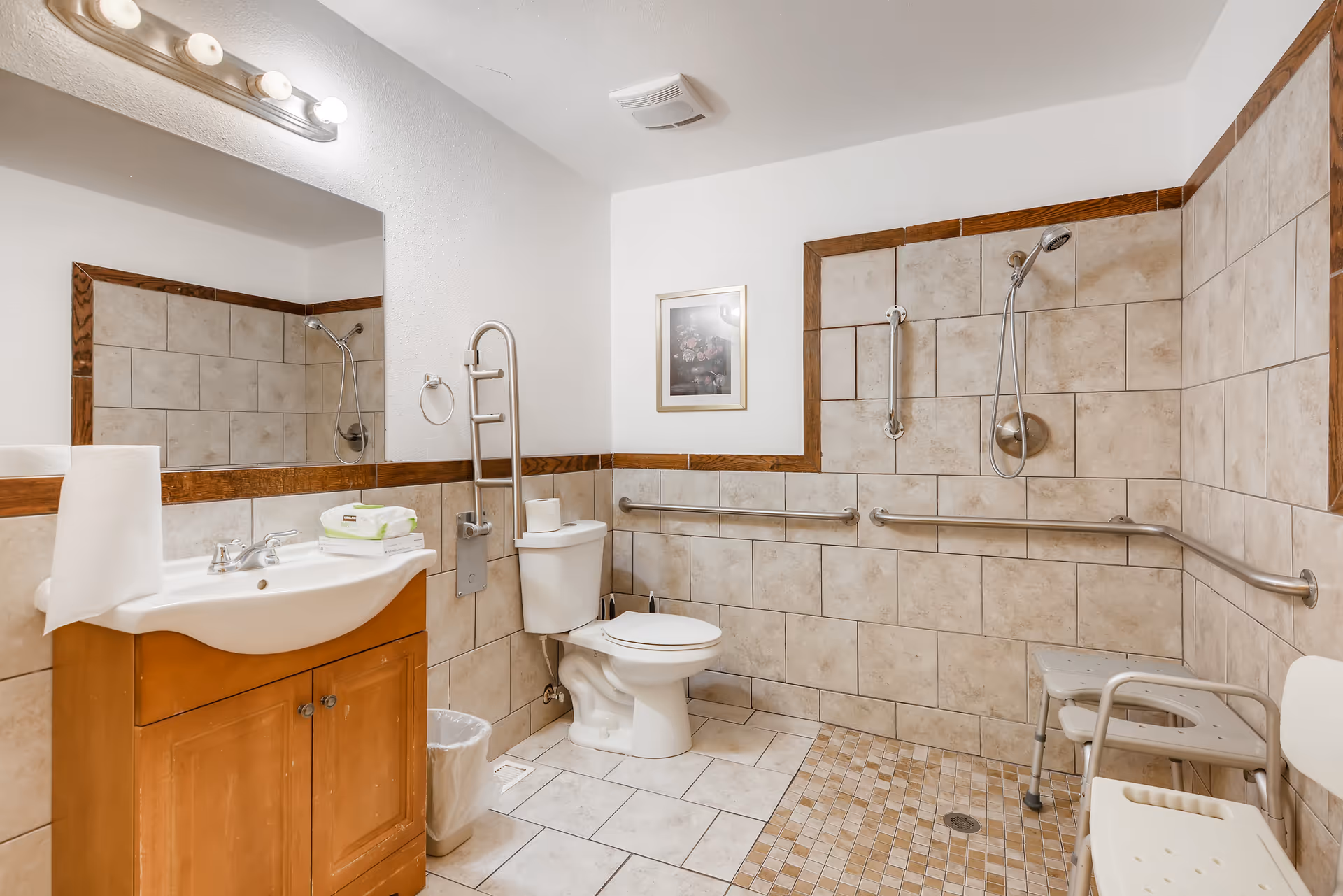 Accessible senior bathroom with a sink and vanity, toilet, multiple grab bars, and a tiled roll-in shower with a shower chair.