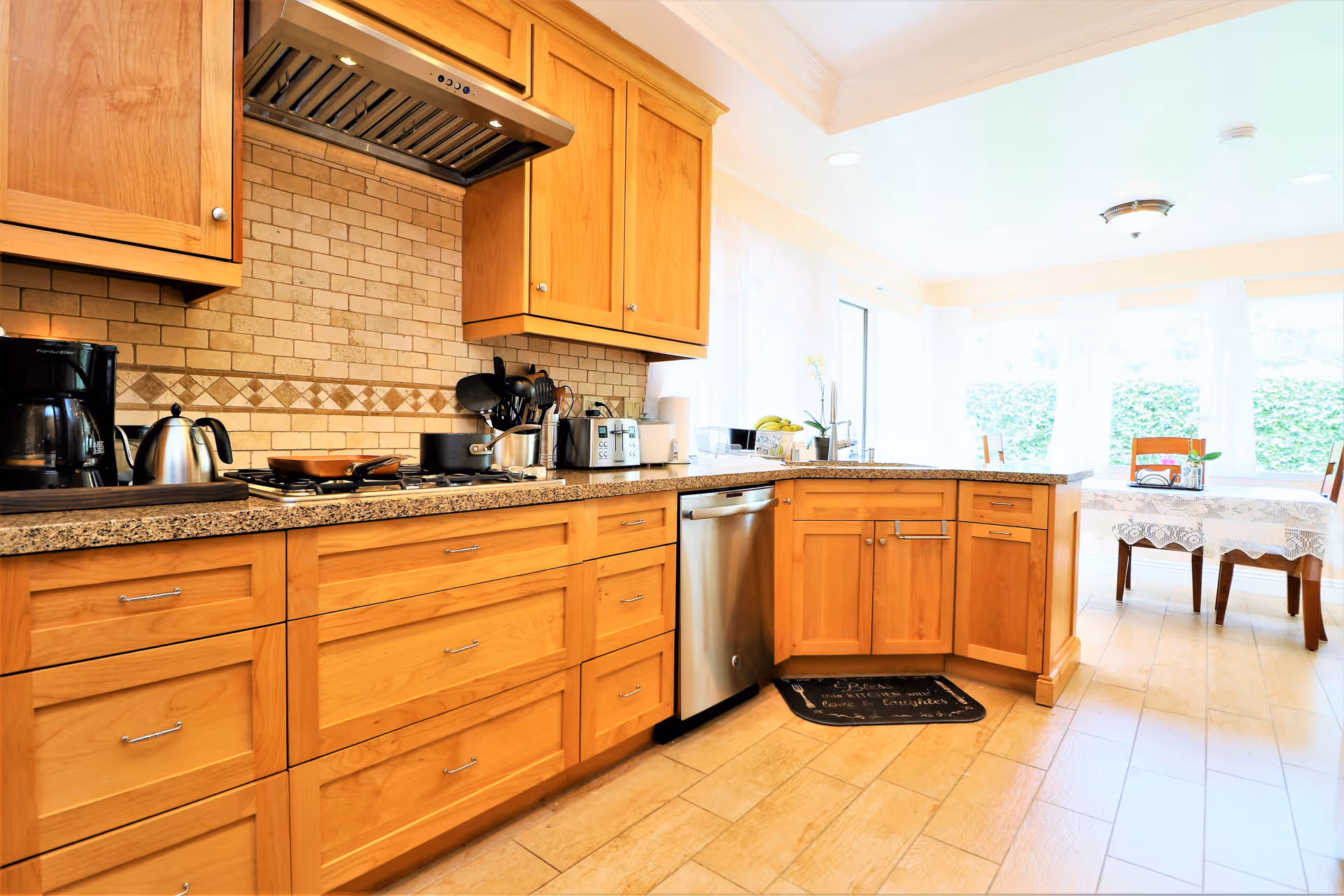 Bright kitchen with wooden cabinets, granite countertops, a stainless steel dishwasher, and various kitchen appliances including a coffee maker, kettle, and toaster. In the background, there is a dining area with a table covered with a lace tablecloth and wooden chairs, next to large windows letting in natural light.