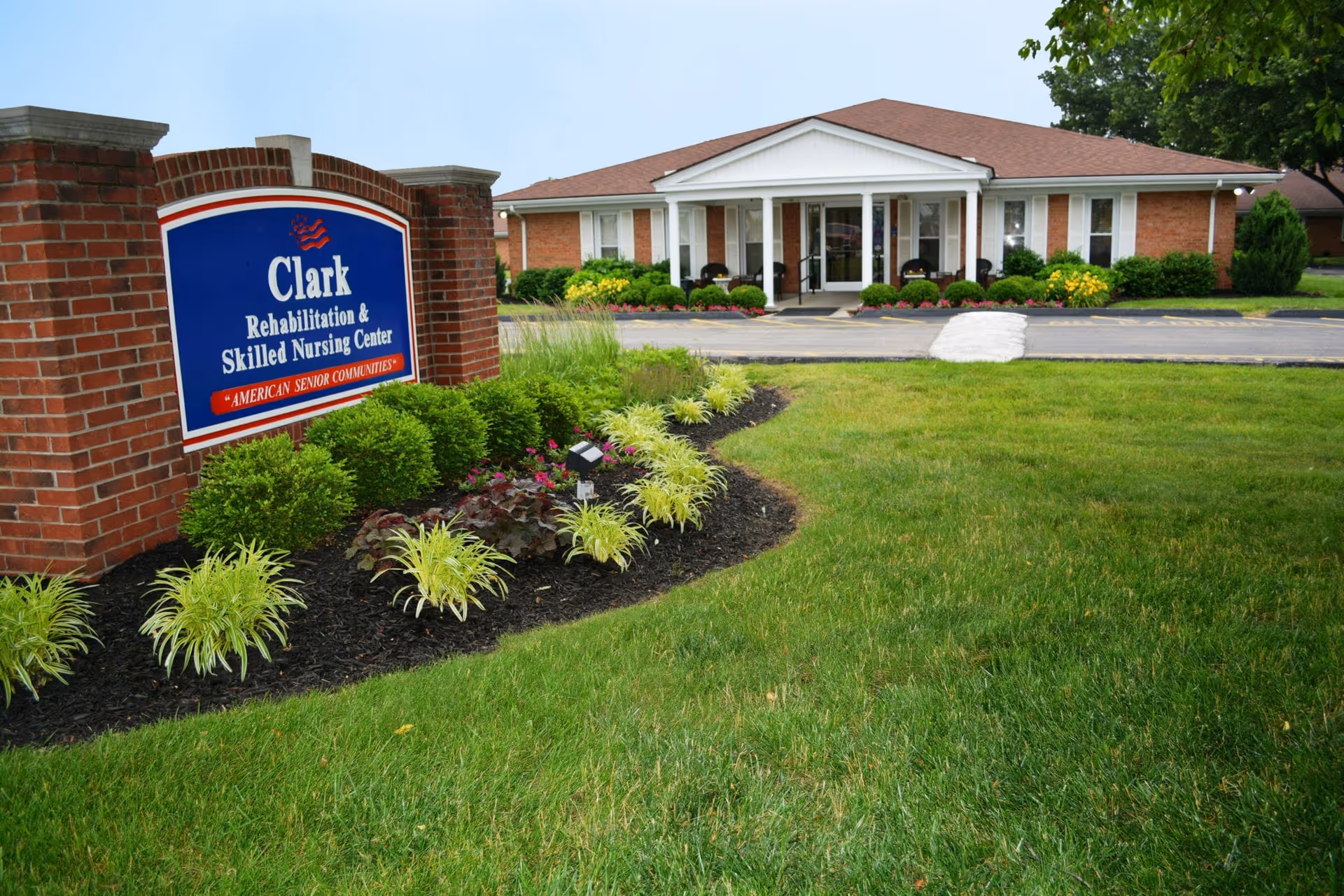 Exterior view of Clark Rehabilitation and Skilled Nursing Center showing a brick sign with the facility name and a single-story building with a porch, surrounded by well-maintained landscaping and green grass.