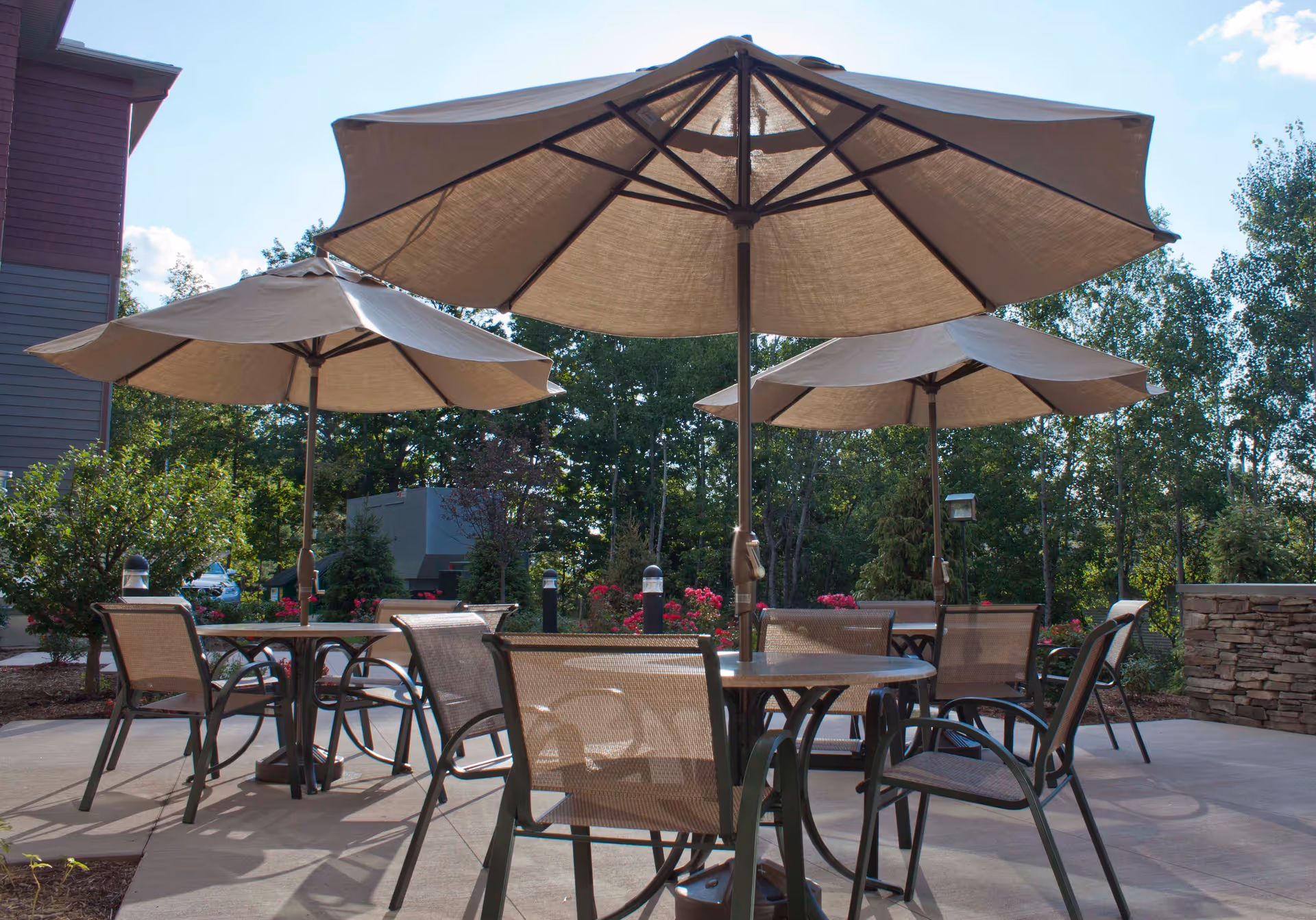 Outdoor patio with round tables, mesh chairs and large beige umbrellas surrounded by trees and landscaping.