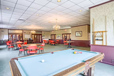 A spacious communal activity room with a pool table in the foreground and multiple round dining tables and chairs under chandeliers.