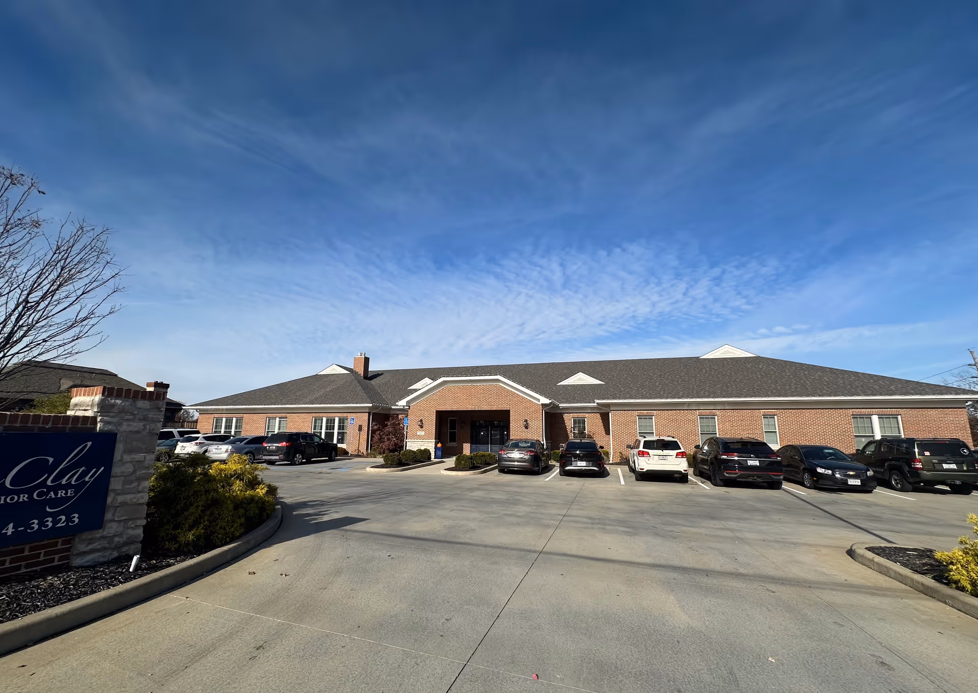 Exterior front view of a single-story brick building with a pitched roof under a partly cloudy blue sky. Several cars are parked in front of the building in a paved parking lot. There is a sign on the left side partially visible with the text 'Clay Senior Care'.