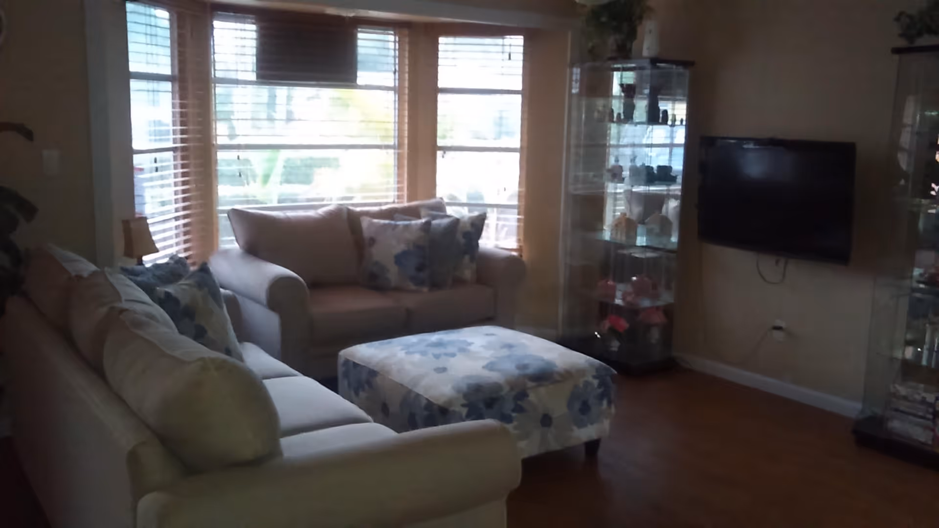 A cozy living room with two beige sofas adorned with floral-patterned cushions, a matching floral ottoman in the center, large windows with blinds letting in natural light, a wall-mounted flat-screen TV, and glass display cabinets filled with decorative items.
