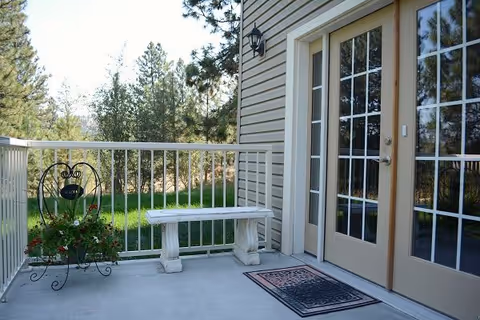 Outdoor patio area with a white railing, a white stone bench, a decorative plant stand with flowers, and double glass doors leading inside. Trees and greenery are visible in the background.